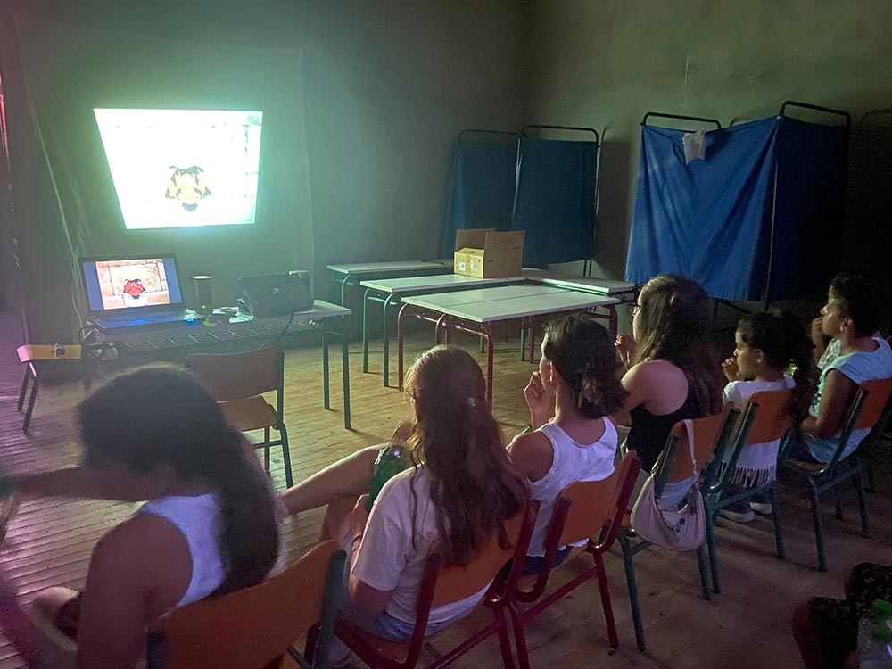 παιδάκια κάθονται στις καρέκλες και βλέπουν ταίνα που προβάλεται στον τοίχο μέσα στο σχολείο